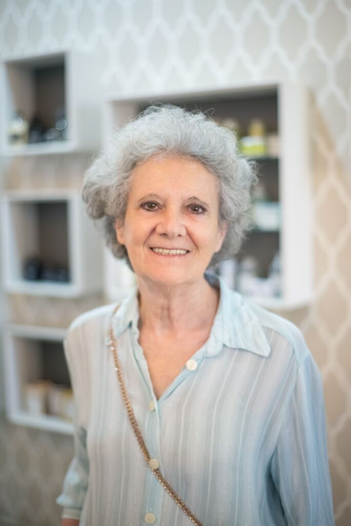 A senior woman with gray hair smiles warmly while standing indoors against a patterned wall.